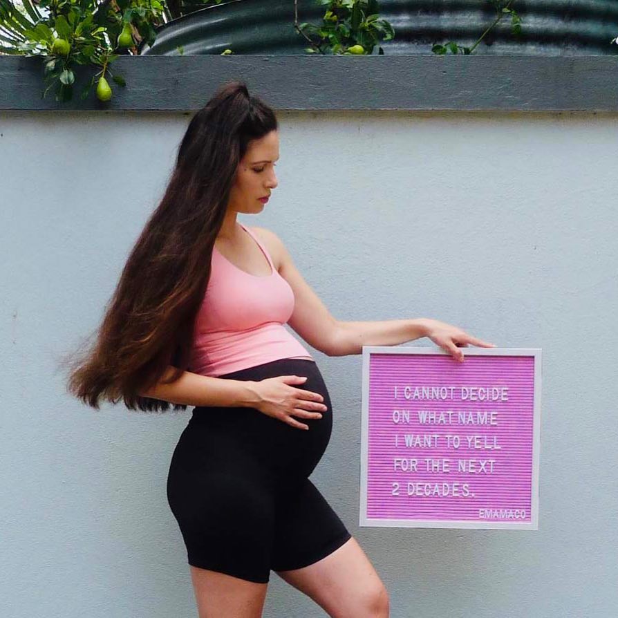 Third trimester pregnant woman on couch in maternity shorts writing a baby name list in a notebook